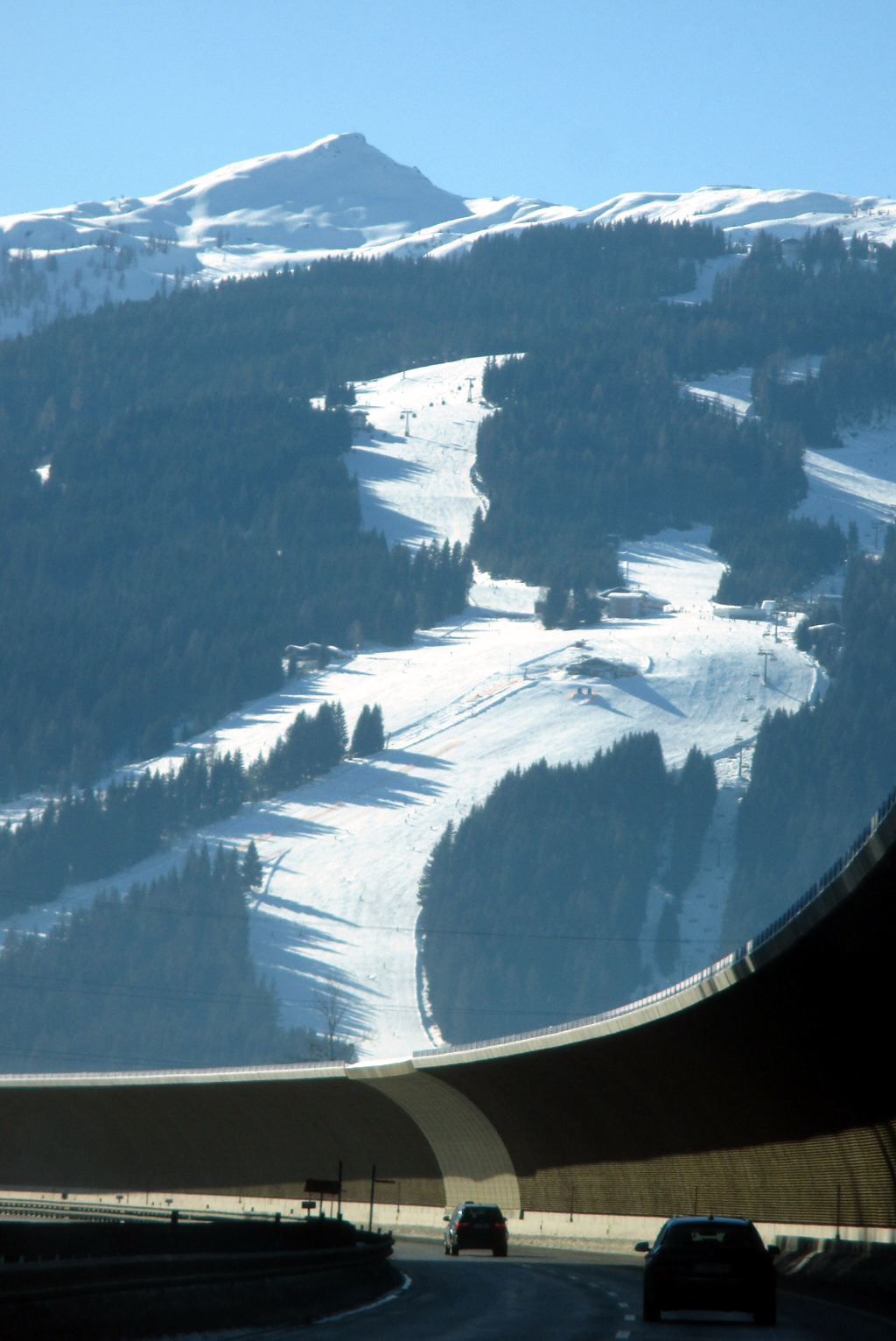 Hinter der Autobahn-Schutzmauer ragen Wälder und im Winter schneebedeckte Berge empor.